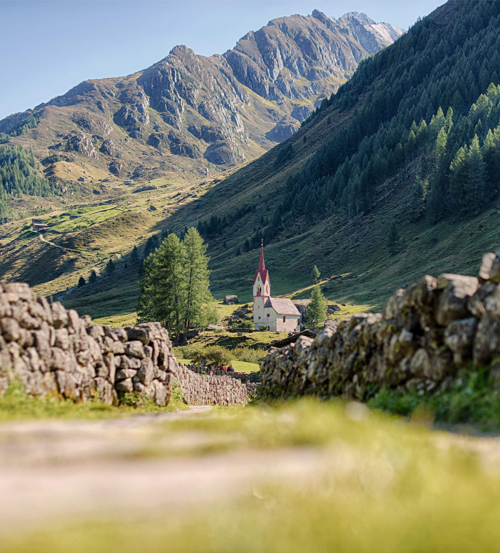 A small church with a red steeple sits in a green mountain valley, surrounded by trees and stone walls. - Ampark