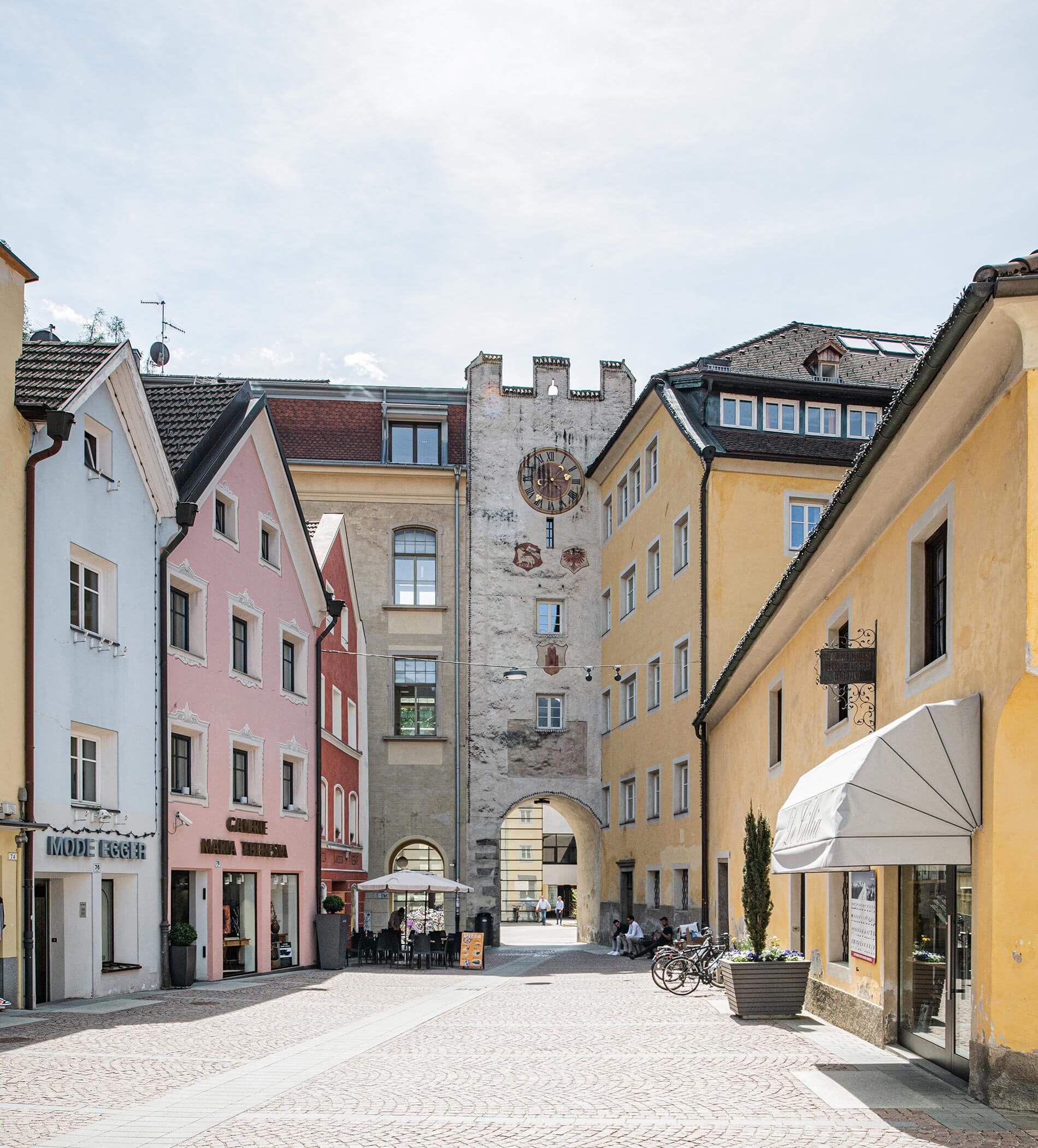 Colorful buildings and a clock tower line a cobblestone street with shops and outdoor seating. - Ampark