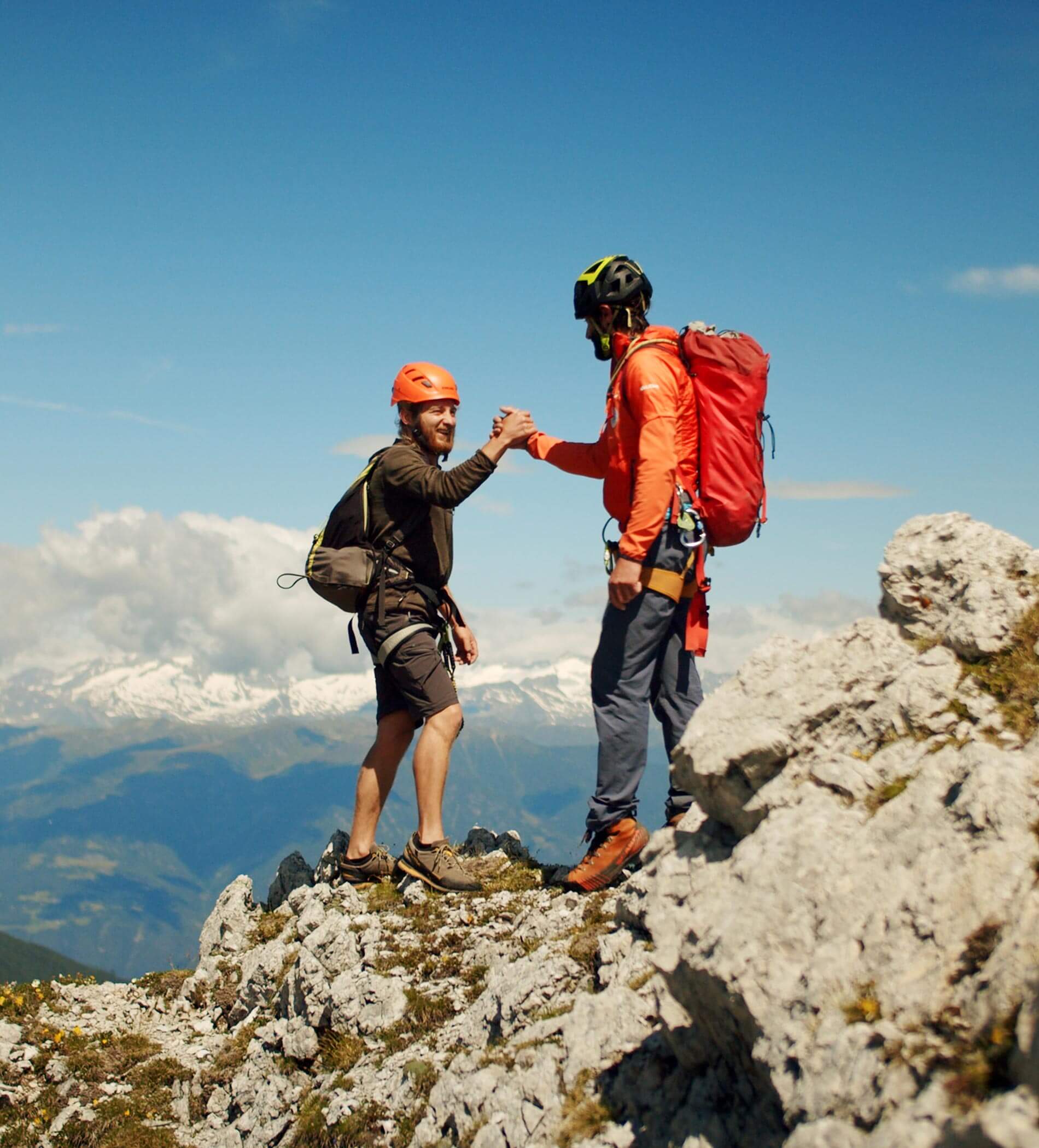 Two hikers in helmets and gear high-five on a rocky mountaintop with snowy peaks in the background. - Ampark