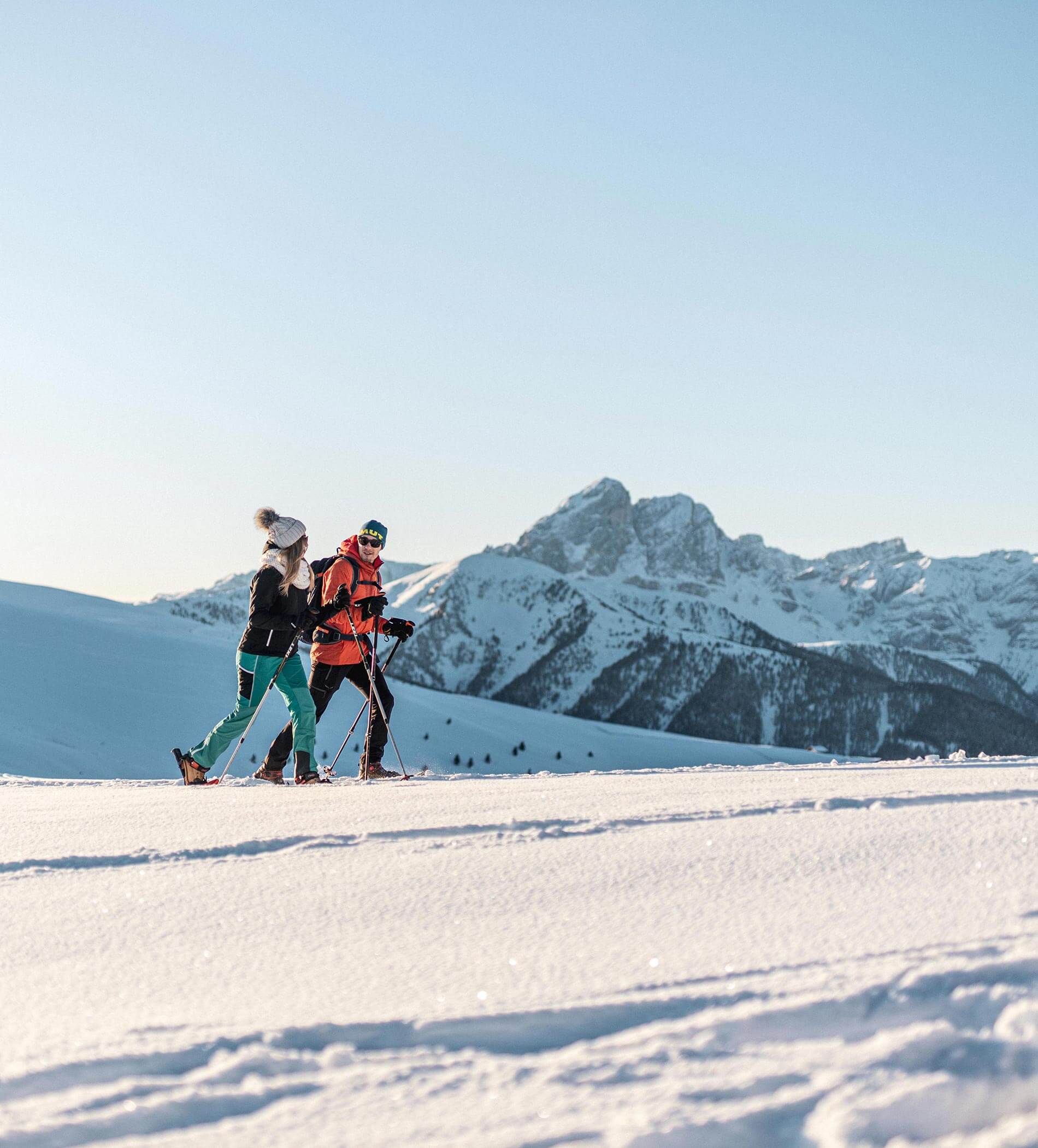 Two people walk with ski poles on snowy terrain, mountains and clear sky in the background. - Ampark