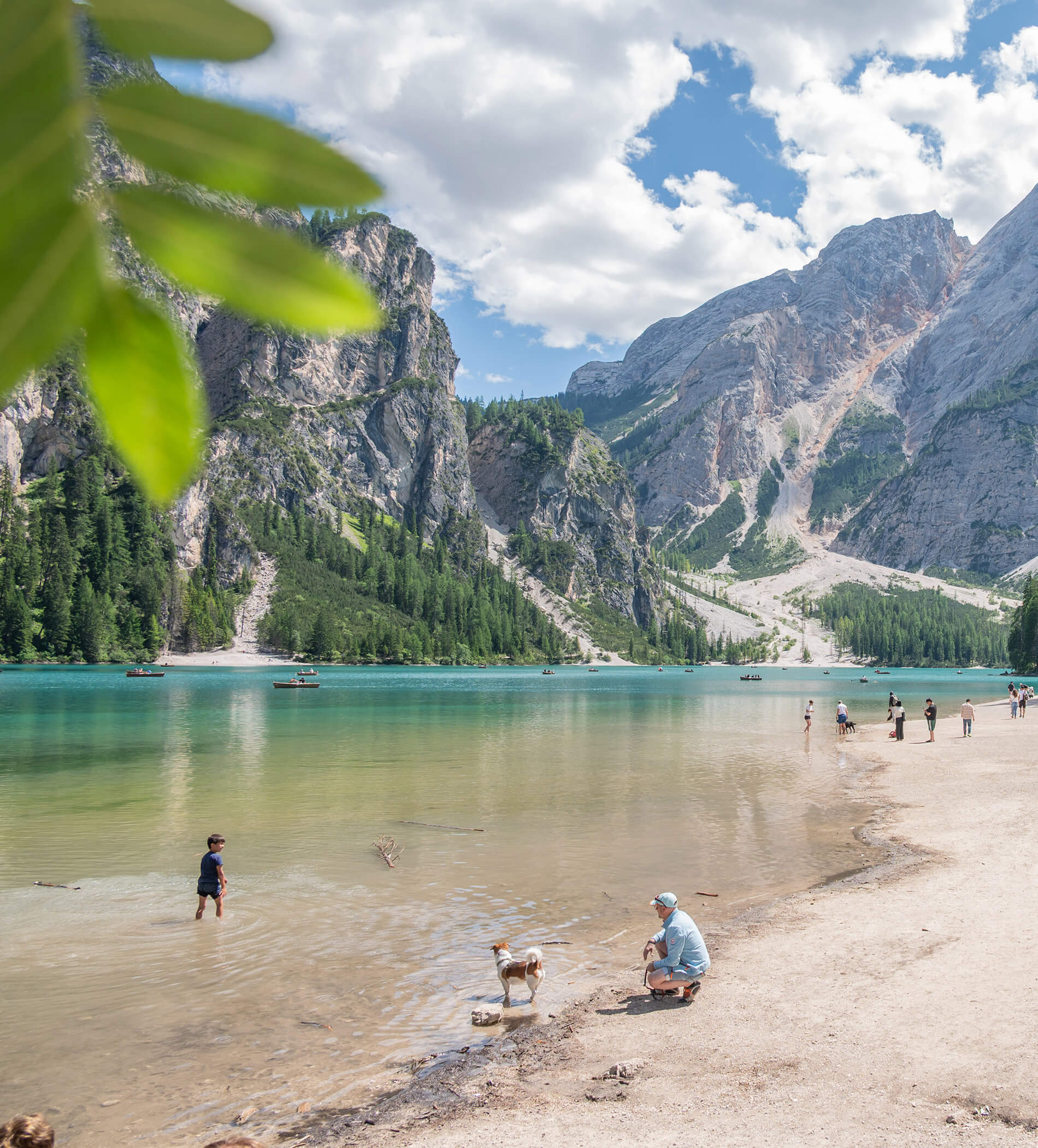 People and a dog relax by a turquoise lake surrounded by mountains, trees, and cloudy blue sky. - Ampark