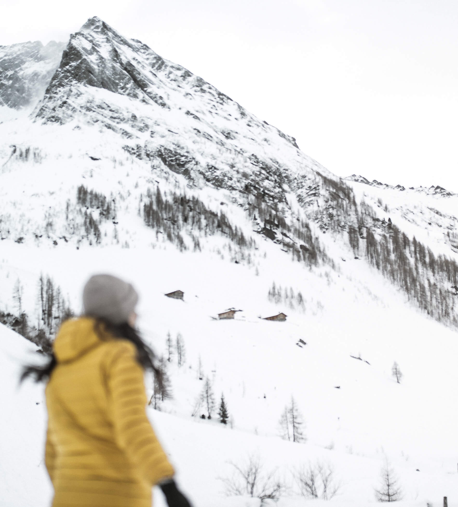 Person in yellow jacket stands in snowy landscape, facing snow-covered mountain with scattered cabins. - Ampark
