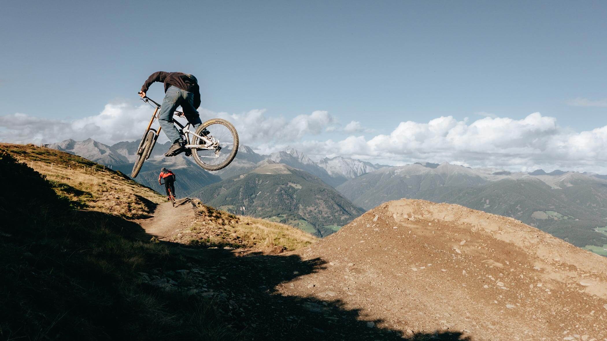 A cyclist jumps off a dirt ramp on a mountain trail with scenic peaks in the background. - Ampark