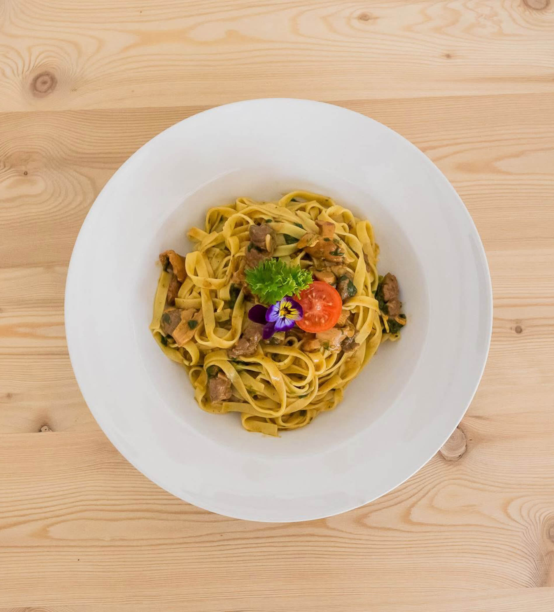 Ein weißer Teller mit Fettuccine, Fleisch, Petersilie, Kirschtomaten und einer essbaren Blüte auf einem Holztisch. - Ampark