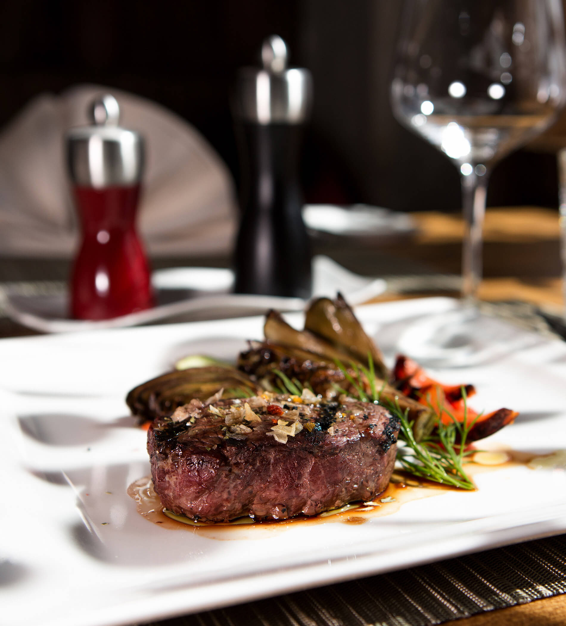 Ein mit Kräutern garniertes Steak auf einem weißen Teller, daneben geröstetes Gemüse und leere Weingläser. - Ampark