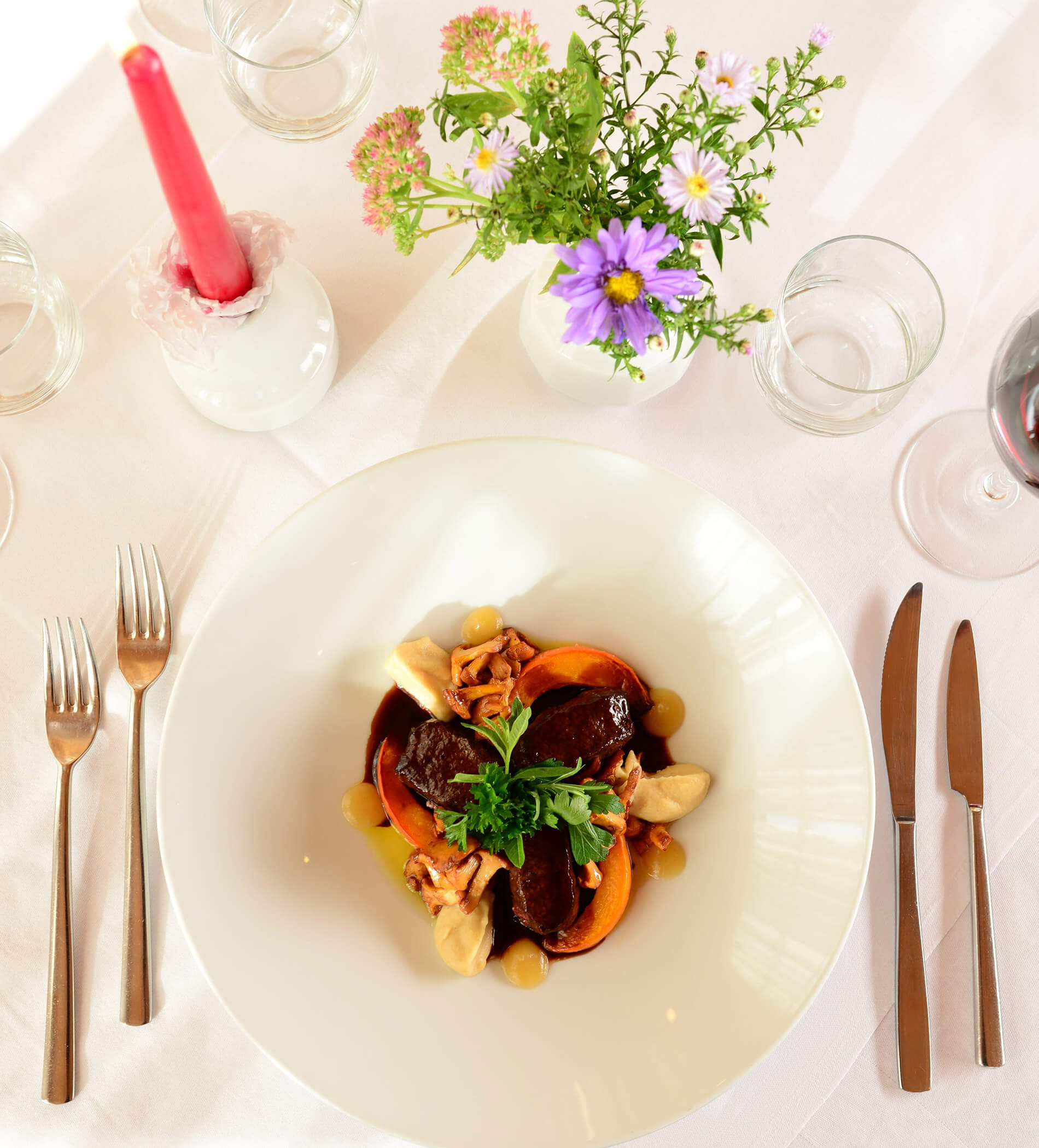 Ein elegant angerichtetes Gericht mit Rindfleisch, Gemüse und Blattgemüse auf einem weißen Tisch, der mit Blumen, einer Kerze und Besteck gedeckt ist. - Ampark