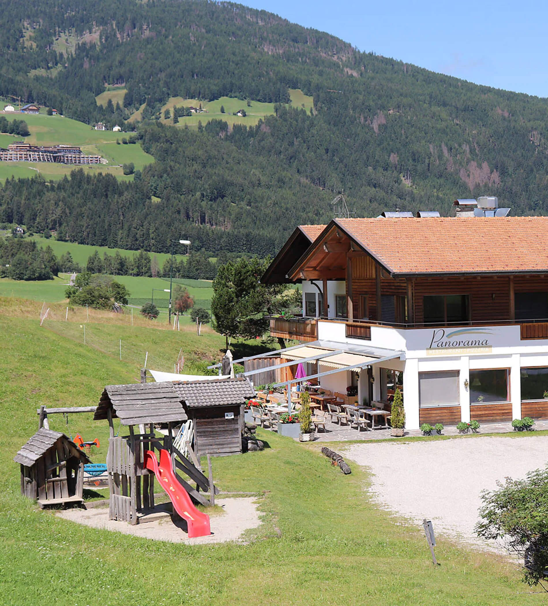 Alpine Pension mit Spielplatz, umgeben von grünen Hügeln und Kiefern unter strahlend blauem Himmel. - Ampark