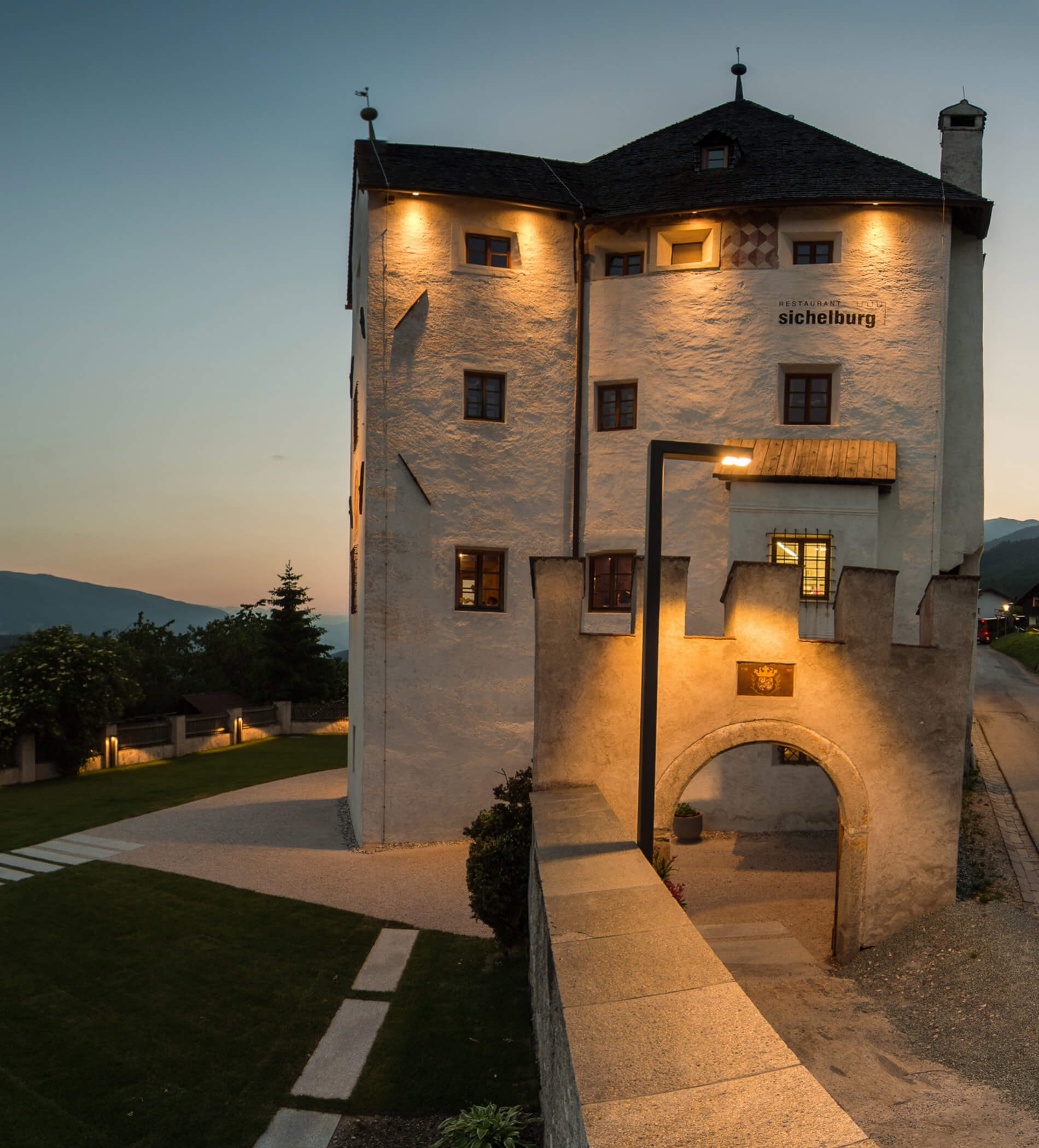 Ein hohes, beleuchtetes Hotel aus Stein mit einem gewölbten Eingang in der Abenddämmerung, neben einer Rasenfläche und mit Blick auf die Berge. - Ampark