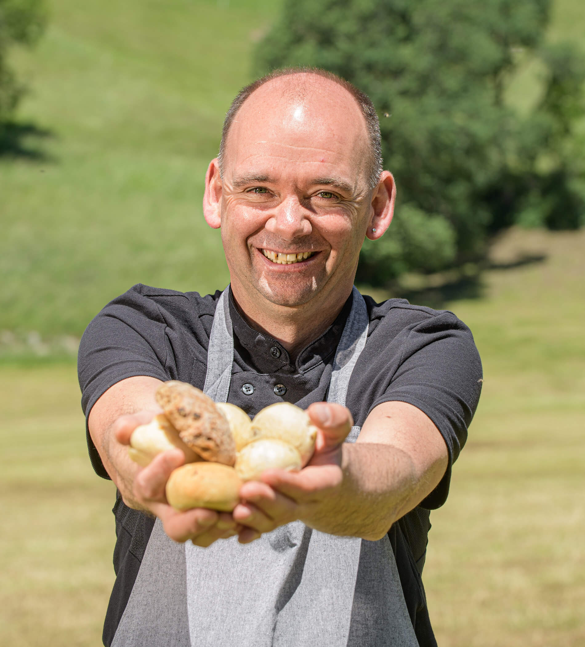 Ein lächelnder Mann in einer Schürze hält auf einem sonnigen Feld mit beiden Händen mehrere Kartoffeln hoch. - Ampark
