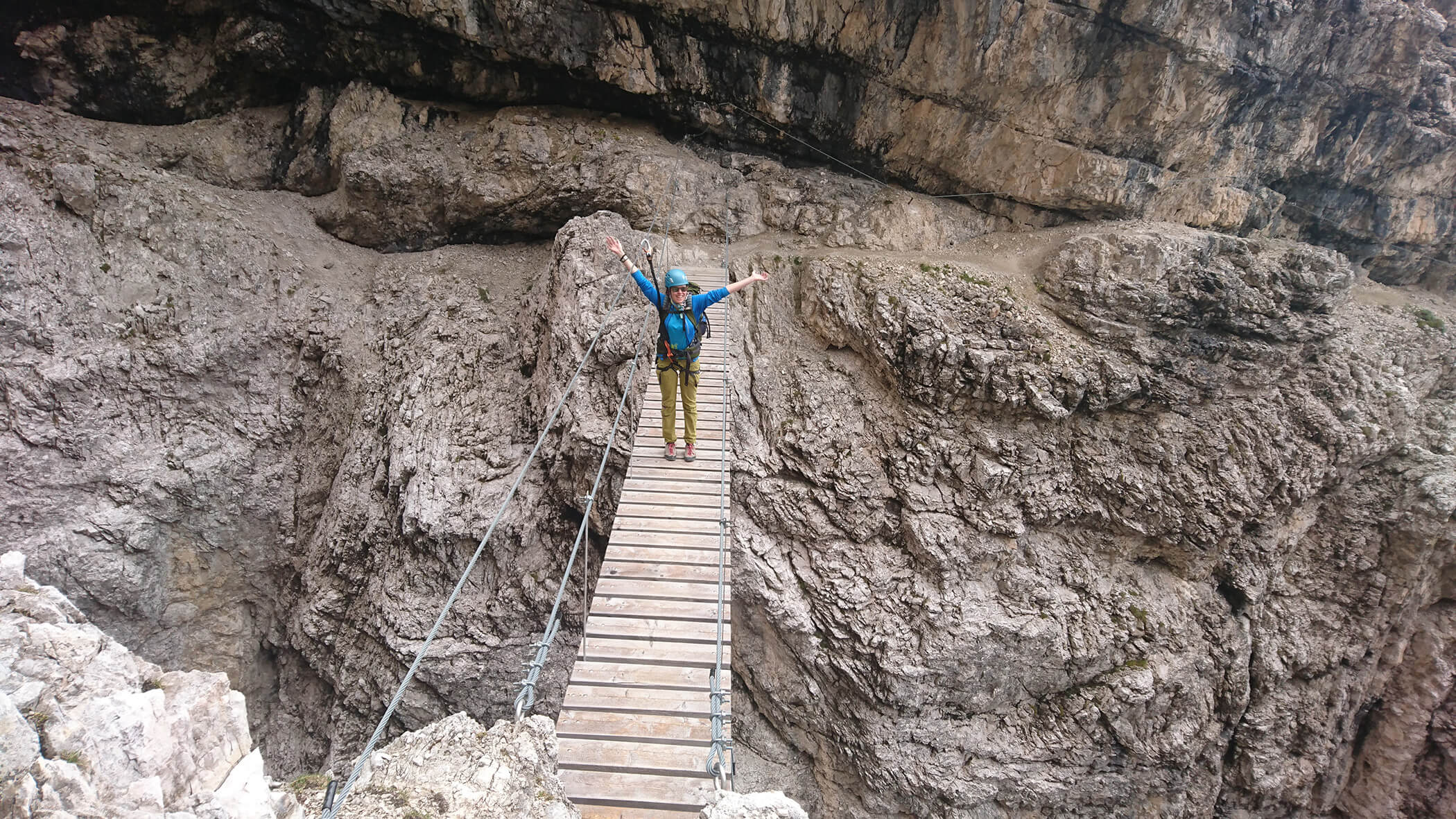Person with raised arms stands on a narrow wooden bridge over rocky terrain in a mountainous area. - Ampark