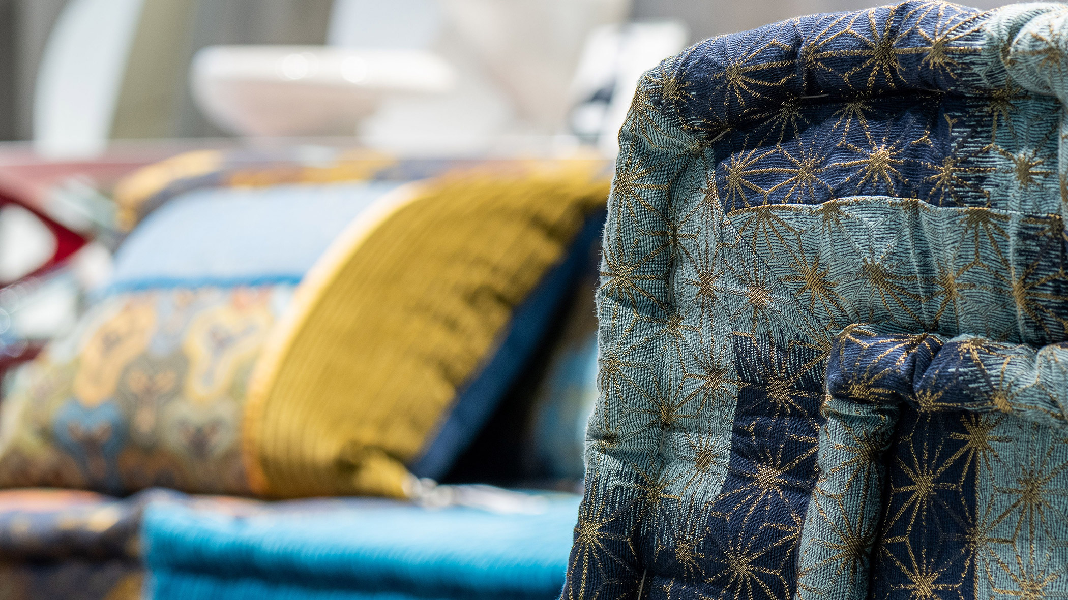 Close-up of a blue and gold patterned quilt, with colorful pillows blurred in the background. - Ampark