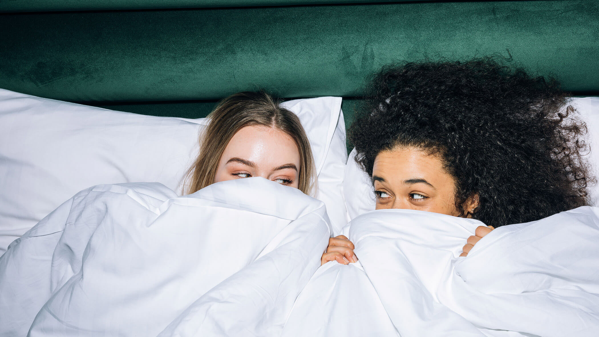 Zwei Frauen liegen im Bett, größtenteils von weißen Laken bedeckt, sehen sich an und lächeln. - Ampark