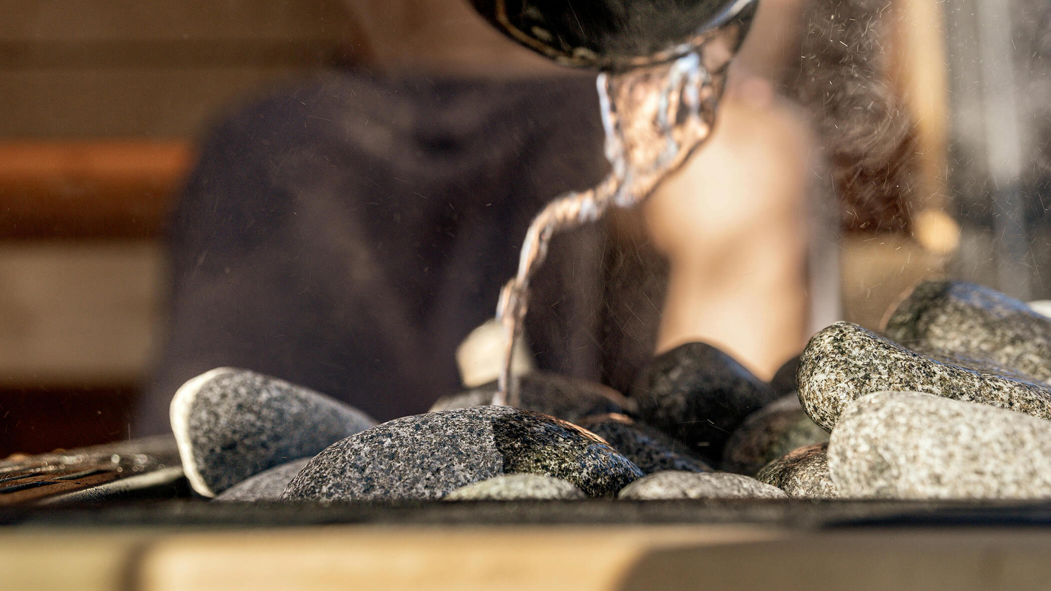 Wasser wird auf heiße Saunasteine gegossen, wodurch Dampf entsteht; im Hintergrund ist eine verschwommene Gestalt zu sehen. - Ampark