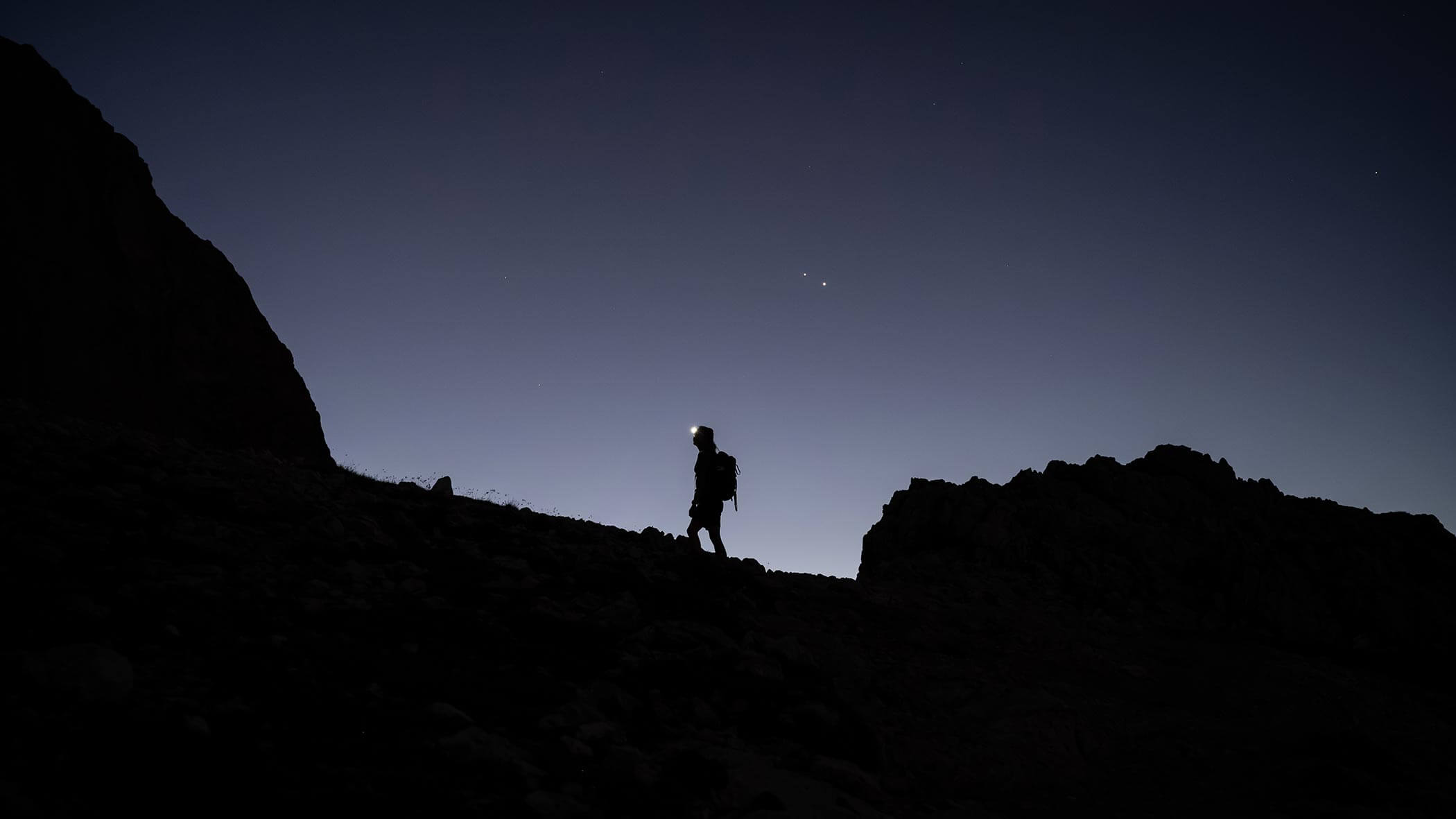 Eine Person mit einer Stirnlampe wandert in der Abenddämmerung bergauf und zeichnet sich als Silhouette vor dem sich verdunkelnden Himmel und dem felsigen Gelände ab. - Ampark