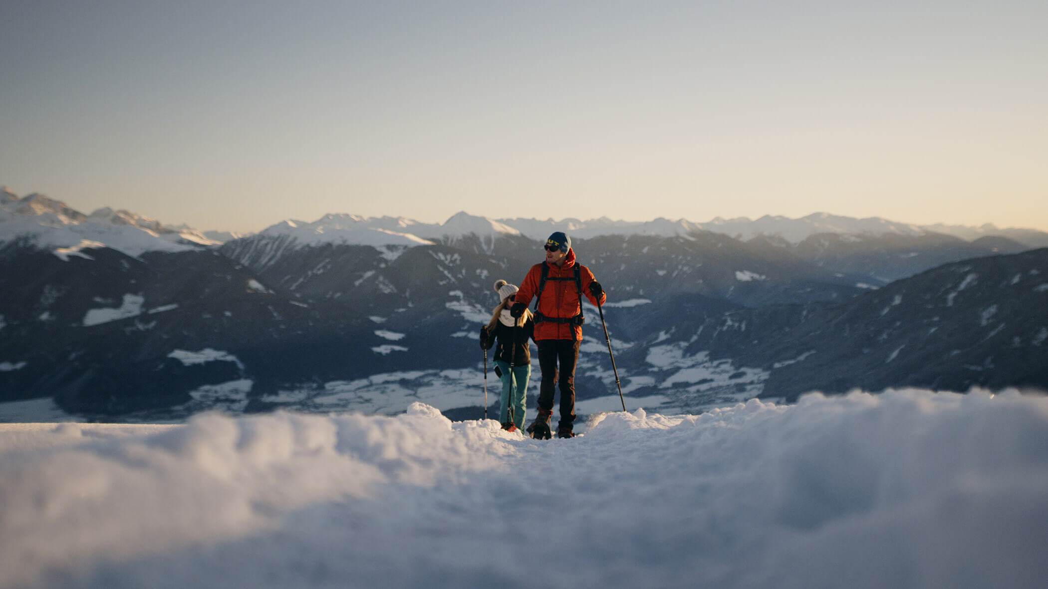 Zwei Menschen wandern bei Sonnenuntergang durch tiefen Schnee, im Hintergrund sind Berge zu sehen. - Ampark