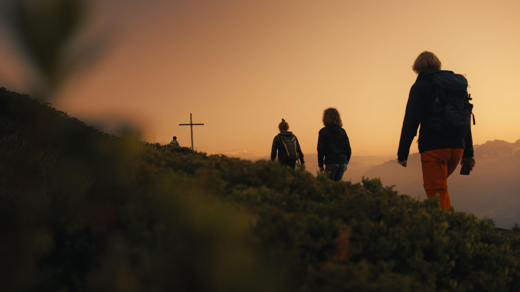 Drei Menschen wandern bei Sonnenuntergang einen grasbewachsenen Hügel hinauf in Richtung eines Kreuzes, im Hintergrund sind Berge zu sehen. - Ampark