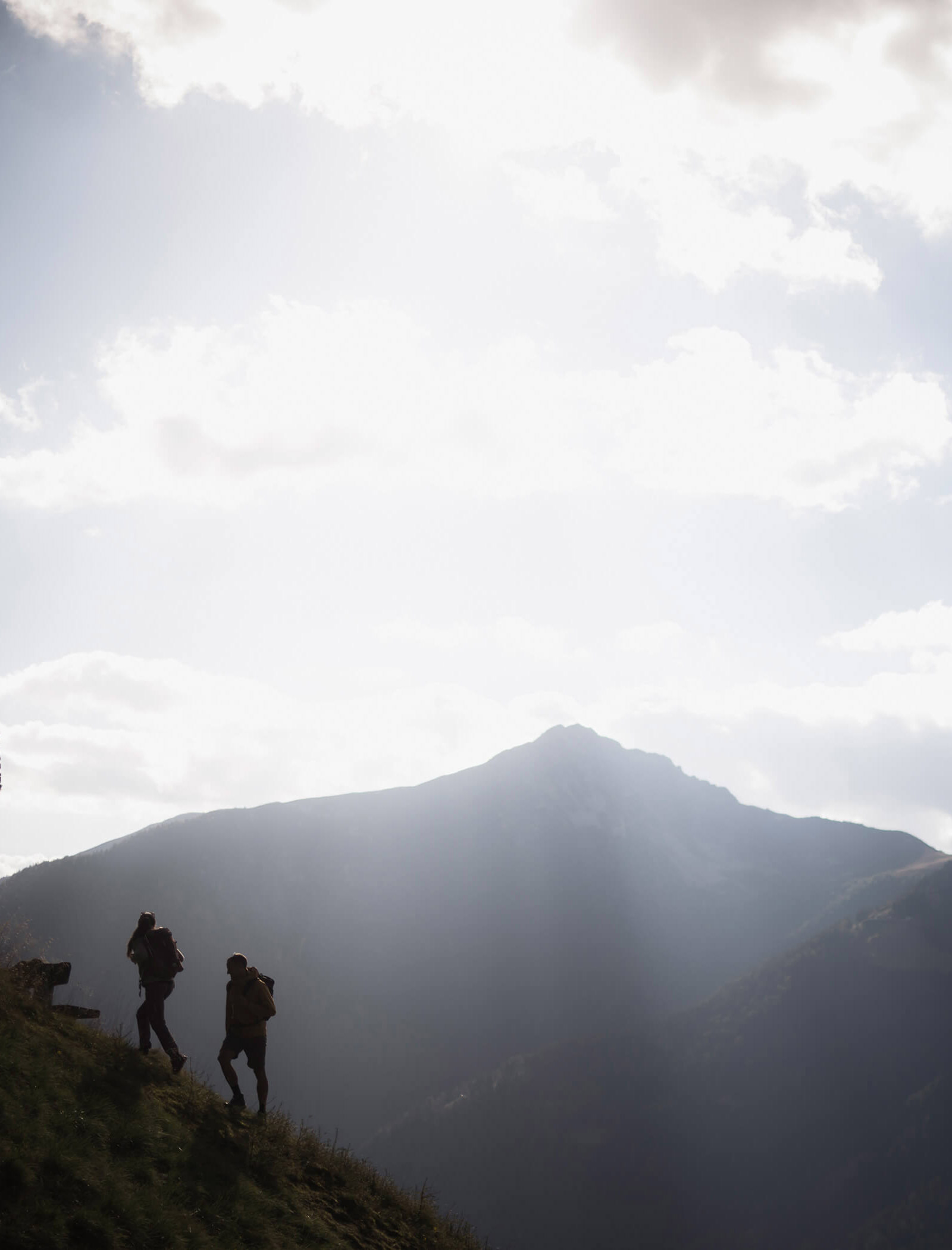 Due escursionisti con gli zaini in spalla salgono su una collina erbosa, con una montagna illuminata dal sole e un cielo nuvoloso sullo sfondo. - Ampark