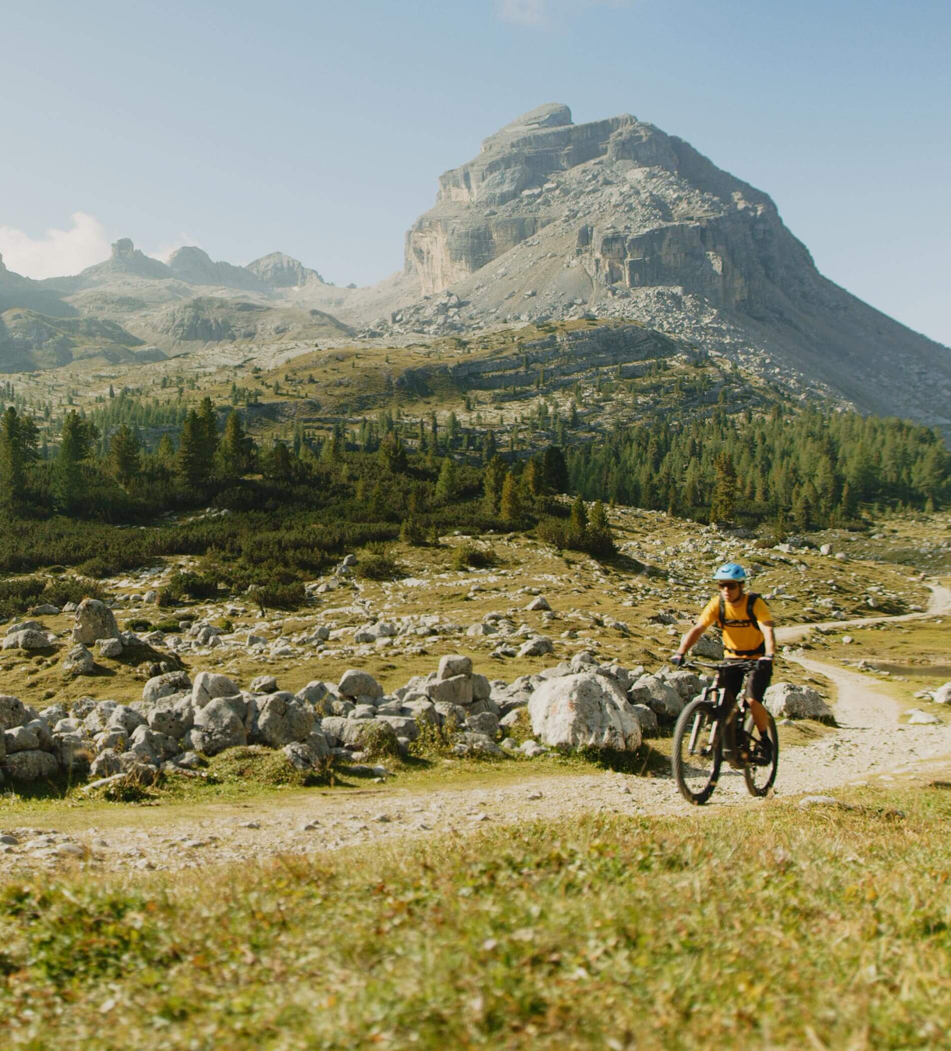 Una persona in bicicletta su un sentiero roccioso, con montagne e alberi sullo sfondo, sotto un cielo limpido. - Ampark
