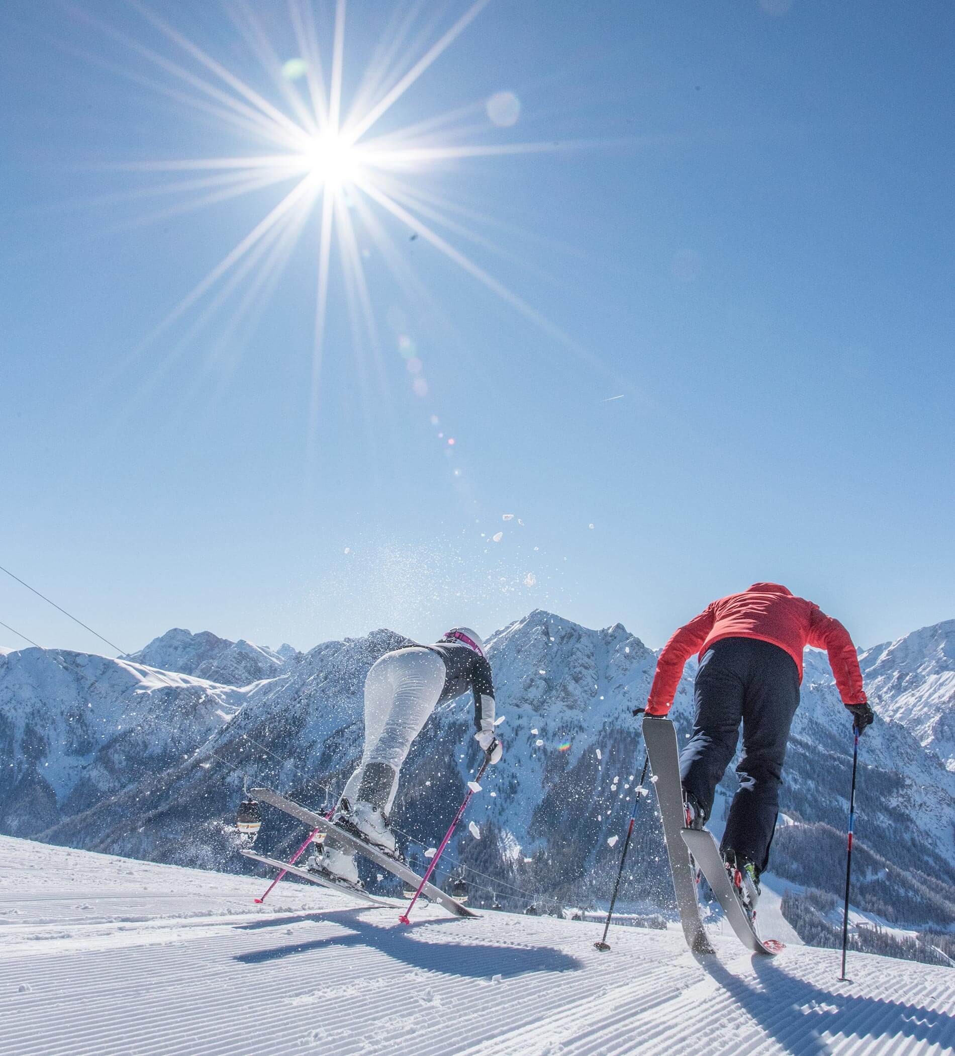 Zwei Skifahrer lassen sich unter strahlendem Sonnenschein auf einem verschneiten Hang mit Bergen im Hintergrund ins Gleiten kommen. - Ampark