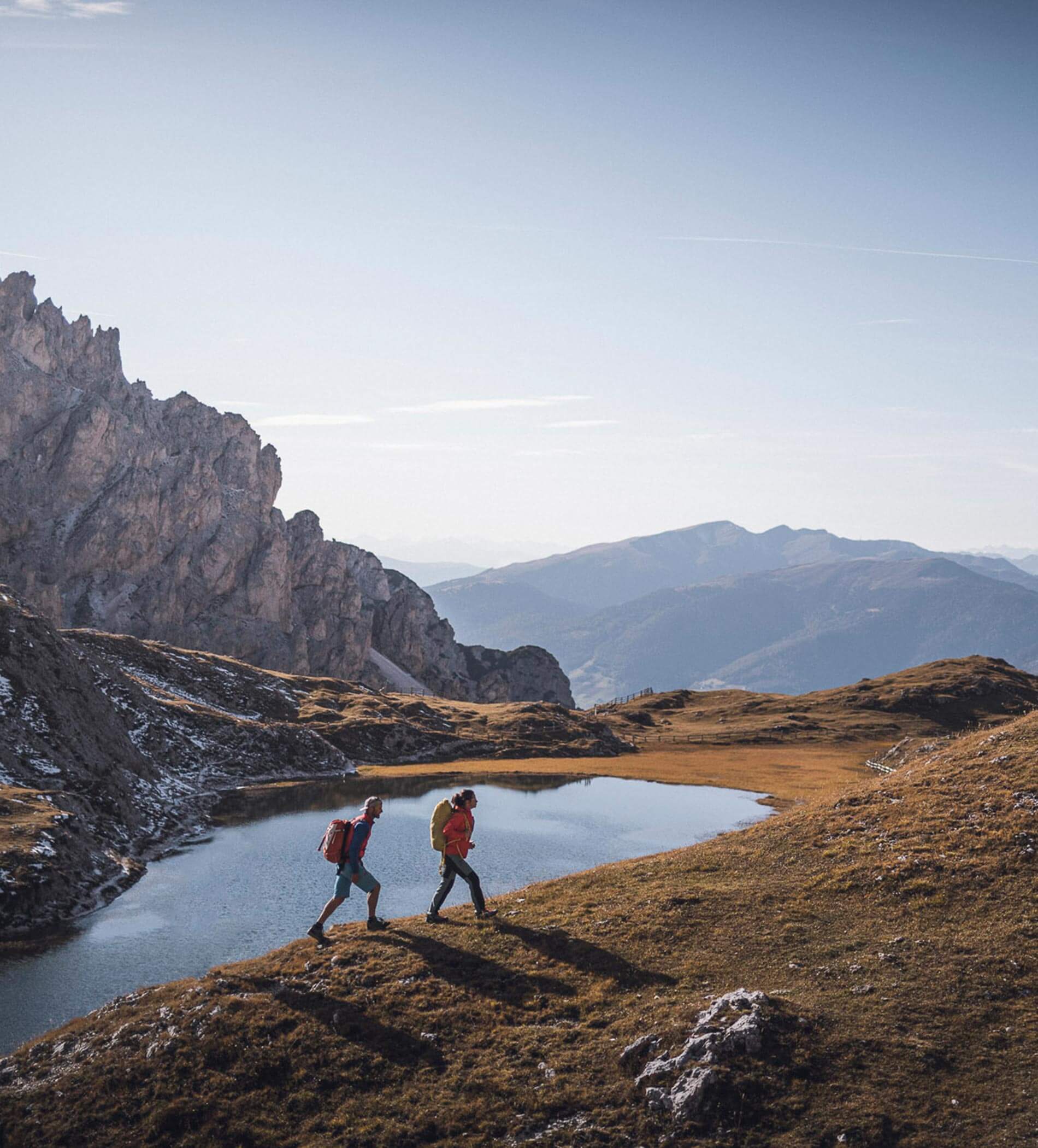 Due escursionisti con gli zaini in spalla camminano lungo le rive di un lago di montagna sotto un cielo limpido, circondati da cime rocciose. - Ampark