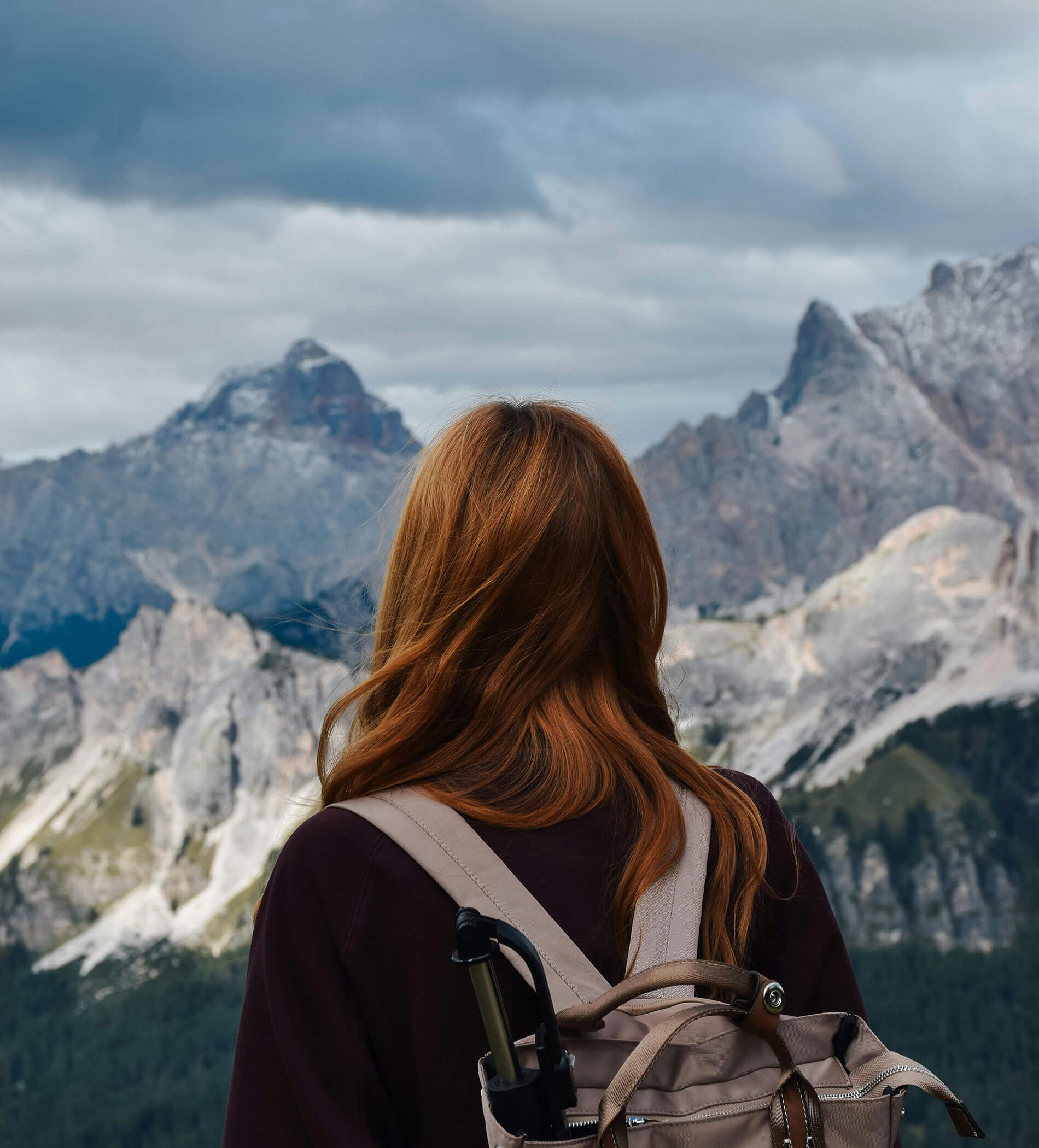 Eine Person mit langen roten Haaren und einem Rucksack blickt unter einem bewölkten Himmel auf die fernen Berge. - Ampark