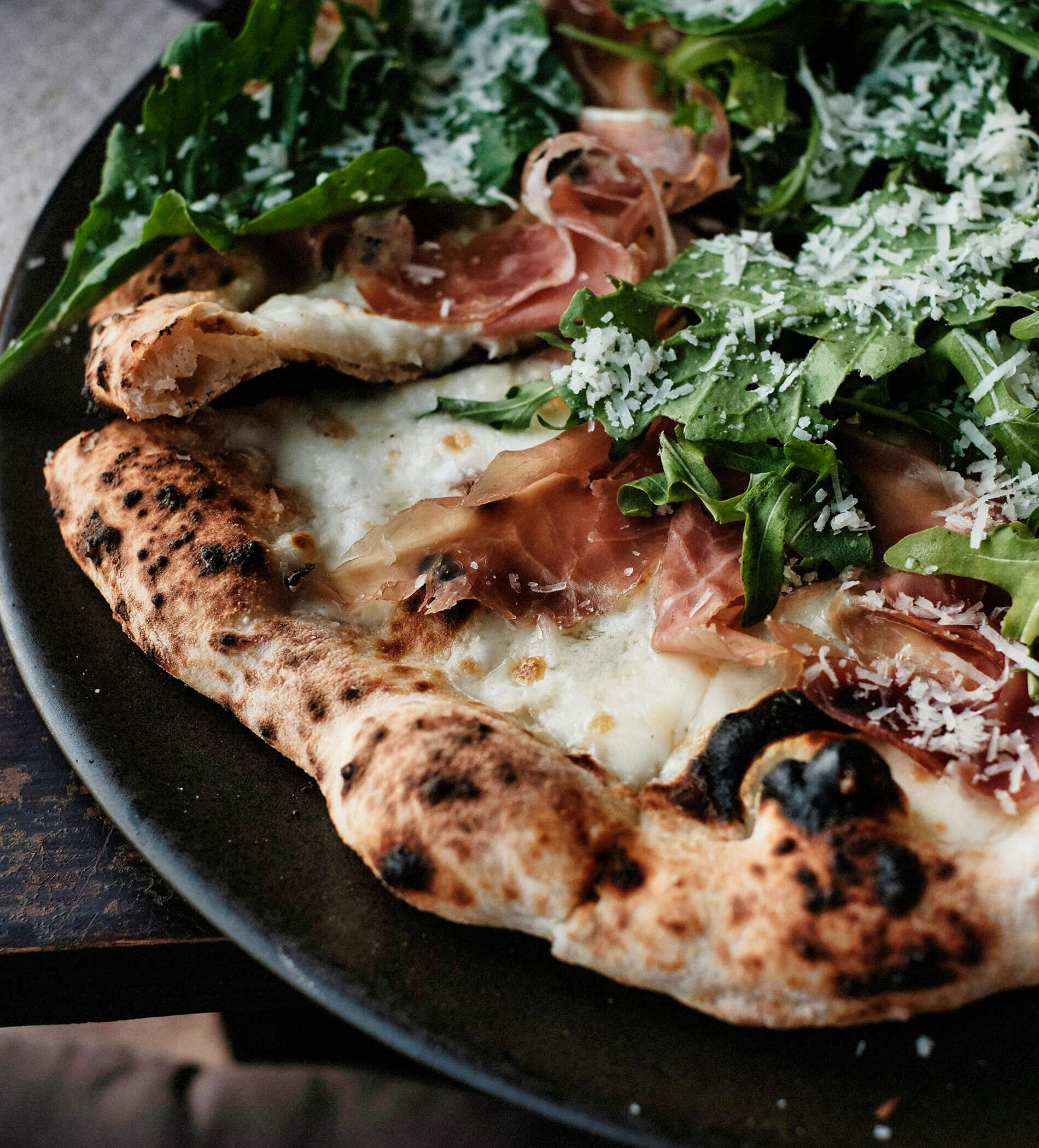 Nahaufnahme einer Pizza mit Rucola, Prosciutto und geriebenem Käse auf einem dunklen Teller. - Ampark