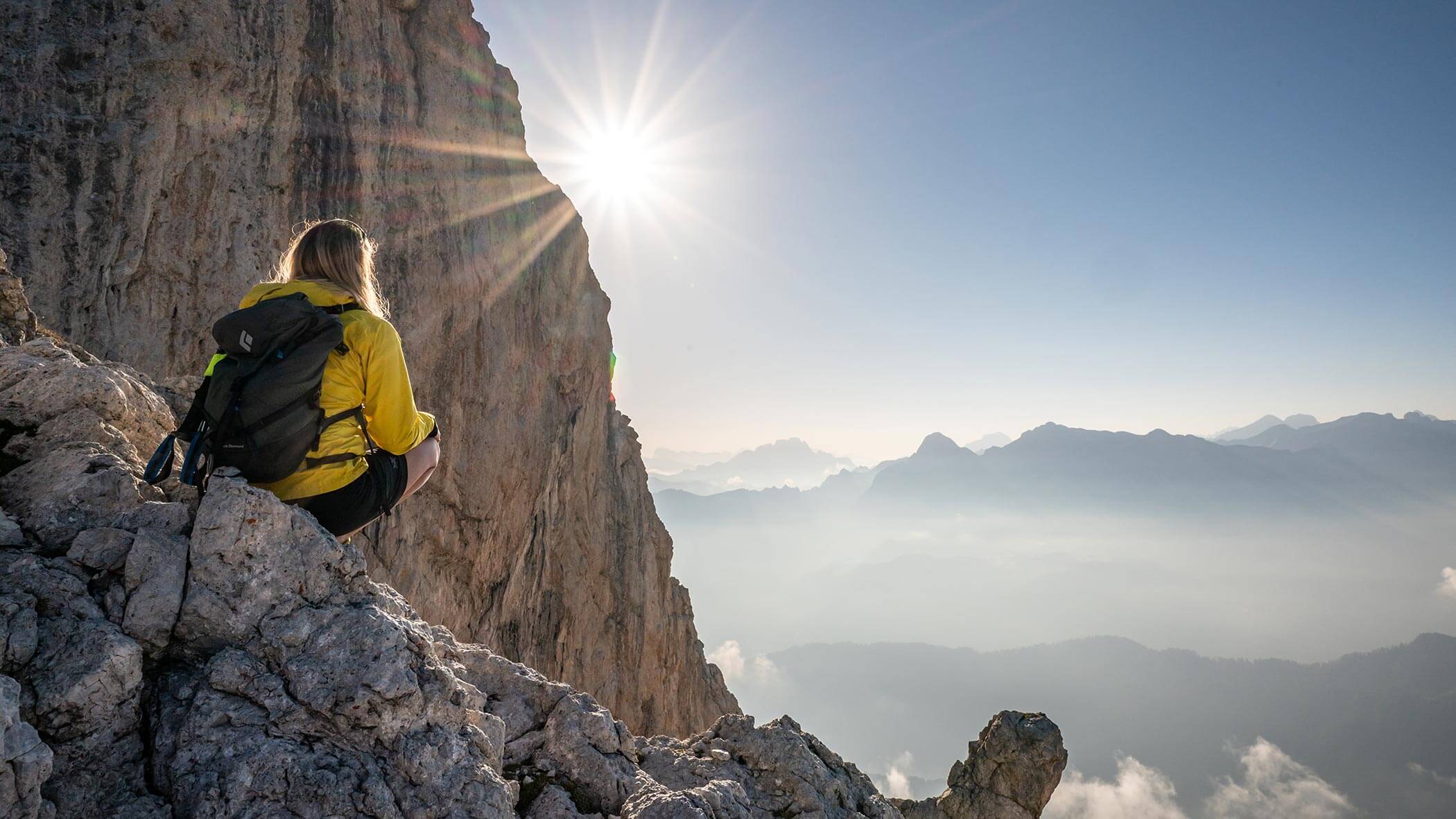 Eine Person in einer gelben Jacke sitzt auf einem felsigen Berghang, im Hintergrund sind der Sonnenaufgang und nebelverhangene Gipfel zu sehen. - Ampark